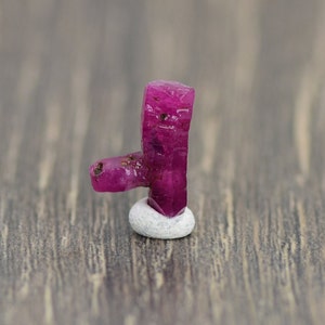 May include: A deep pink ruby crystal with a rough, faceted surface. The crystal is on a small white pedestal.