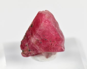 May include: A deep pink, faceted, rough-cut gemstone with black inclusions. The gemstone is sitting on a white surface.