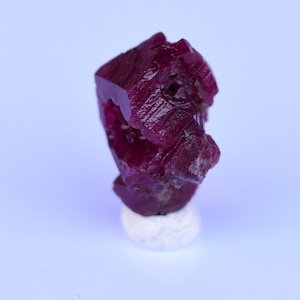 Puede incluir: Un cristal de rubí rojo intenso con una forma geométrica facetada. El cristal se muestra sobre un pedestal blanco sobre un fondo morado claro.