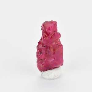 May include: A deep red ruby crystal with a rough, natural texture. The crystal is on a white display stand.