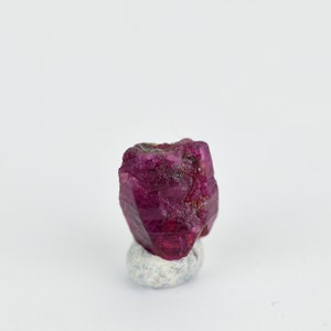 May include: A deep red ruby crystal with a faceted, rough-cut shape. The crystal is sitting on a small, white, textured base.