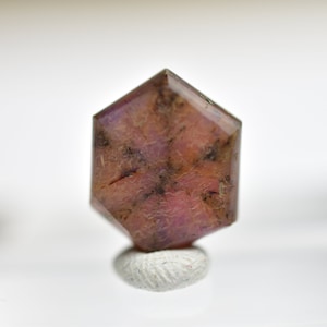 Puede incluir: Una gema hexagonal facetada, de color marrón rojizo, salpicada de inclusiones más oscuras. La gema está montada sobre una pequeña base cilíndrica blanca, sobre un fondo blanco suave.