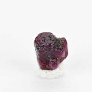 May include: A deep red ruby gemstone with a rough, natural surface. The ruby is sitting on a white, textured surface.