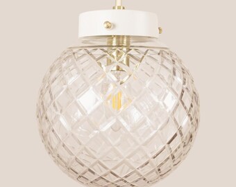 May include: A clear glass globe pendant light with a diamond pattern. The light fixture has a white top and a gold-colored base. The cord is a cream color. The light bulb is visible inside the globe.