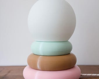 May include: A table lamp featuring a spherical white glass shade resting on a stack of three circular, ring-shaped bases. The base rings are colored in light mint green, tan, and soft pink, arranged in a vertical, tiered, and rounded design.