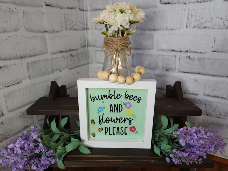May include: A white framed sign with a green background and the text "bumble bees and flowers please" in black script. The sign is decorated with small yellow bees and colorful flowers.