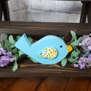 May include: A blue wooden bird with a yellow beak and a floral patterned fabric on its chest. The bird is sitting on a dark brown wooden shelf.