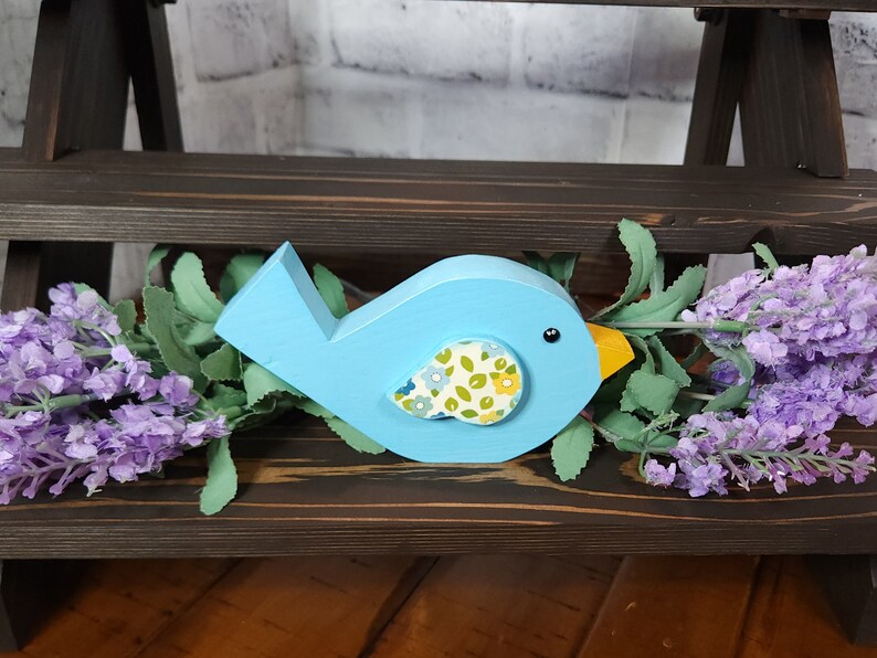 May include: A blue wooden bird with a yellow beak and a floral patterned fabric on its chest. The bird is sitting on a dark brown wooden shelf.