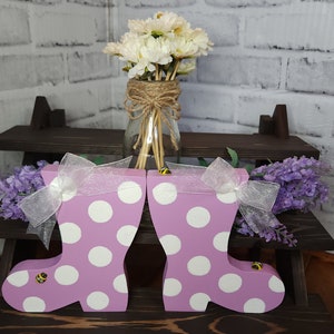 May include: Two pink rain boots with white polka dots and yellow bee accents. The boots are on a wooden shelf with a bouquet of white flowers in a glass jar.
