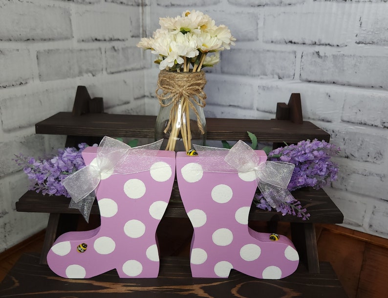 May include: Two pink rain boots with white polka dots and yellow bee accents. The boots are on a wooden shelf with a bouquet of white flowers in a glass jar.
