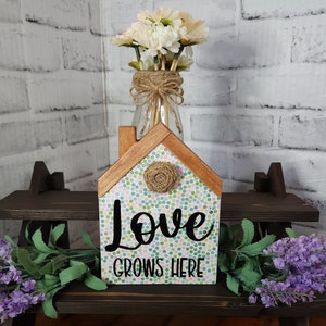 May include: A wooden house-shaped sign with a white background and colorful polka dots. The sign says "Love Grows Here" in black lettering. The sign is sitting on a dark brown shelf with a vase of white flowers and a sprig of lavender.