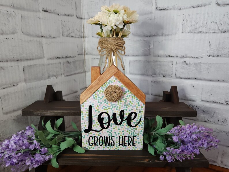 May include: A wooden house-shaped sign with a white background and colorful polka dots. The sign says "Love Grows Here" in black lettering. The sign is sitting on a dark brown shelf with a vase of white flowers and a sprig of lavender.