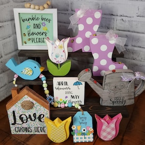 May include: A tiered wooden shelf display featuring a variety of spring-themed wooden decorations. The decorations include a bird, a house, a watering can, a pair of rain boots, tulips, and signs with the text "bumble bees and flowers please", "Love grows here", "April showers bring May flowers", and "Hello Spring".