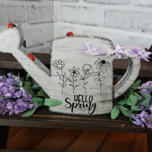 May include: White wooden watering can with a black and white floral design and the text "Hello Spring". The watering can has a purple and white checkered ribbon tied around the handle. There are red ladybug accents on the watering can.