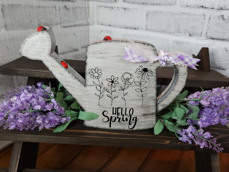 May include: White wooden watering can with a black and white floral design and the text "Hello Spring". The watering can has a purple and white checkered ribbon tied around the handle. There are red ladybug accents on the watering can.