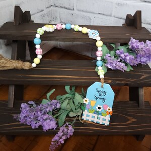 May include: A spring-themed beaded garland with a wooden tag that reads "Spring was Spring". The garland is made of white beads with colorful gingham and flower accents. The tag has a blue background with a white picket fence and colorful flowers.