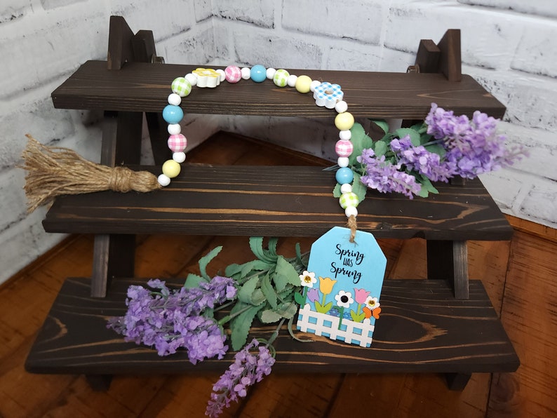 May include: A spring-themed beaded garland with a wooden tag that reads "Spring was Spring". The garland is made of white beads with colorful gingham and flower accents. The tag has a blue background with a white picket fence and colorful flowers.
