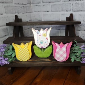 May include: Three wooden tulips with different patterned designs. The tulips are yellow with white gingham, white with floral print, and pink with white gingham. The tulips are displayed on a two-tiered wooden shelf.