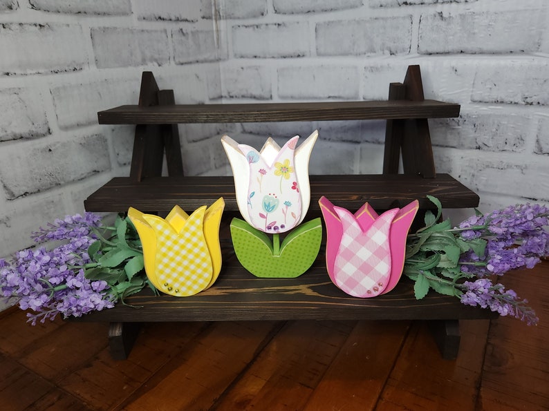 May include: Three wooden tulips with different patterned designs. The tulips are yellow with white gingham, white with floral print, and pink with white gingham. The tulips are displayed on a two-tiered wooden shelf.