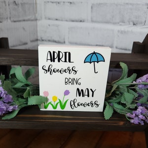 May include: White wooden sign with the text "April showers bring May flowers" in black lettering. A blue umbrella is above the word "bring". Three colorful flowers are below the word "bring".