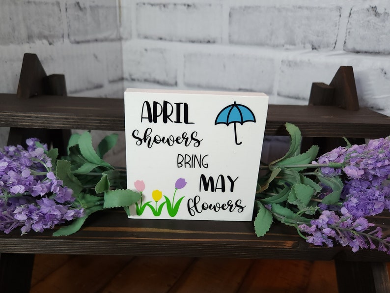 May include: White wooden sign with the text "April showers bring May flowers" in black lettering. A blue umbrella is above the word "bring". Three colorful flowers are below the word "bring".