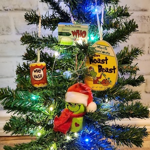 May include: A green Grinch Christmas ornament with a red scarf and a white Santa hat hanging on a green Christmas tree with lights. The tree is decorated with other ornaments, including a yellow and red can that says "Who Hash" and a yellow and red can that says "Roast Beast".