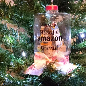May include: A clear glass ornament with a red ribbon and a white bow. The ornament has a handwritten message that says "stressed, blessed & amazon obsessed".