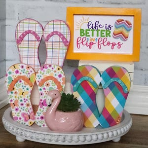 May include: A white tray with a beaded edge, decorated with four pairs of colorful flip flops and a pink flamingo figurine. A sign above the flip flops reads "Life is better in flip flops".
