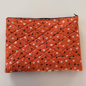 May include: An orange quilted pouch with a black zipper. The fabric is patterned with white ghosts, black witch hats, and orange pumpkins. The pouch has a diamond pattern.