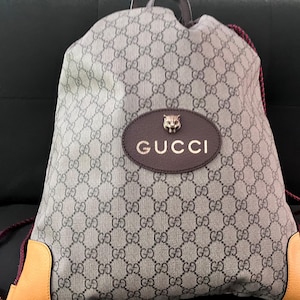 Peut inclure: Un sac à dos à cordon avec un motif monogramme gris et noir. Le centre présente un écusson ovale marron foncé avec le mot GUCCI en lettres dorées et un petit emblème de tête de tigre en métal au-dessus. La base a des coins en cuir beige.