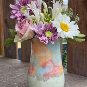 Puede incluir: Un jarrón de cerámica con una base lisa y redondeada, con un esmalte multicolor que presenta tonos mezclados de verde pálido, naranja suave y azul claro. El jarrón contiene un arreglo floral de margaritas blancas, rosas rosadas y crisantemos rayados.