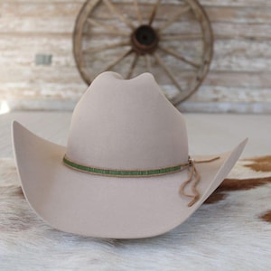 May include: A light beige cowboy hat with a green beaded hatband and a leather chin strap. The hat has a classic western design with a curved brim and a tall crown. The background includes a wooden wagon wheel and a rustic wooden wall.
