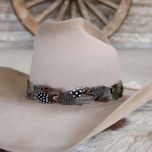 May include: A light beige felt cowboy hat featuring a wide brim and a high crown. The base of the crown is encircled by a decorative band composed of layered, multi-patterned feathers in shades of brown, black, and white with distinct spots.