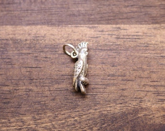 May include: A small, silver-toned metal charm shaped like a cockatoo bird. The charm features detailed engravings of feathers and a crest on the head, with a circular jump ring attached at the top for hanging on a necklace or bracelet chain.