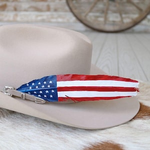 May include: A tan cowboy hat with a decorative feather painted with the American flag design. The feather is blue with white stars and red and white stripes. The hat has a leather band with a silver buckle.