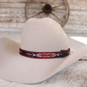 May include: A light beige cowboy hat with a decorative band featuring a geometric pattern in red, black, and turquoise. The hat has a classic western design, suitable for outdoor wear.