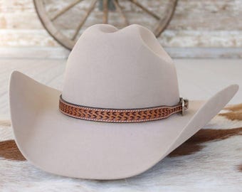 May include: A light beige cowboy hat with a brown leather band featuring a decorative pattern. The hat has a classic western design with a curved brim and a tall crown. The hat is displayed on a cowhide rug.