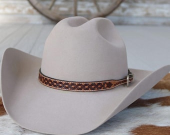 May include: A light beige cowboy hat with a decorative brown leather band. The hat has a classic western design with a curved brim and a tall crown. The leather band features a repeating circular pattern.