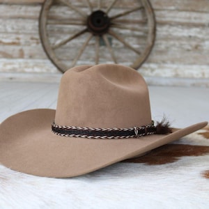 May include: A tan cowboy hat with a wide brim and a decorative band. The band is made of woven brown and white material. A small feather is attached to the side of the hat. The hat is displayed on a cowhide rug.