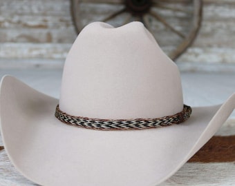 May include: A light beige cowboy hat with a decorative woven band in brown, black, and white. The hat has a classic western design with a curved brim and a tall crown. The background features a rustic wooden wall and a wagon wheel.