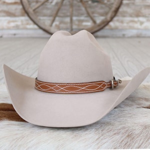 May include: A light beige felt cowboy hat featuring a structured crown and a wide, curved brim. A tan leather hat band wraps around the base, decorated with white stitched geometric patterns and a small silver-toned metal buckle closure.