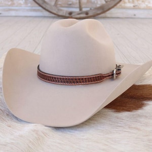 May include: A beige cowboy hat with a brown leather band featuring a woven pattern. The hat has a classic western design with a curved brim and a rounded crown. The hat is displayed on a cowhide rug.