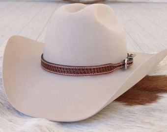 May include: A beige cowboy hat with a brown leather band featuring a woven pattern. The hat has a classic western design with a curved brim and a rounded crown. The hat is displayed on a cowhide rug.