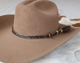 May include: A brown felt cowboy hat with a braided leather hatband and a long brown leather hat string.
