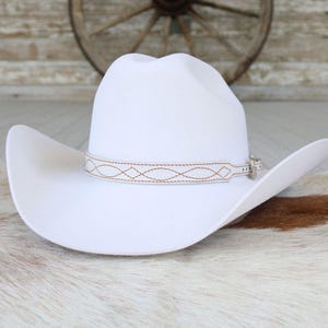 May include: A solid white cowboy hat featuring a wide brim and a creased crown. The hat is accented with a thin, light-colored hat band that displays a repeating geometric diamond pattern stitched in a contrasting brown thread and a metal buckle.