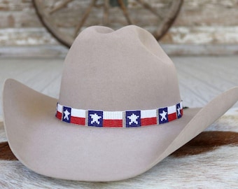 May include: A light beige felt cowboy hat featuring a beaded hatband. The hatband displays a repeating pattern of the Texas state flag, consisting of a white star on a blue square next to a horizontal red and white stripe design.