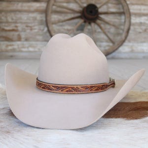 May include: A light beige cowboy hat with a brown leather band featuring an embossed floral design. The hat has a wide brim and a classic western style. The background includes a wooden wall and a wagon wheel.