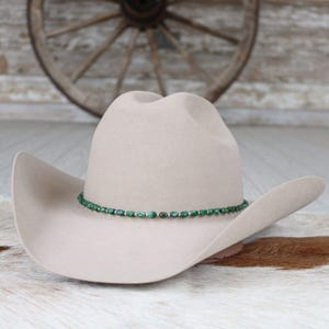 May include: A light tan cowboy hat with a decorative green beaded band. The hat has a wide brim and a classic western design. The hat is displayed on a cowhide rug.