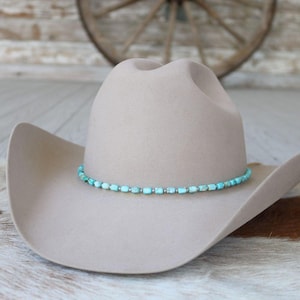 May include: A beige cowboy hat with a turquoise beaded hatband. The hat is made of a soft material and has a classic western design. The turquoise beads are small and evenly spaced around the hat.