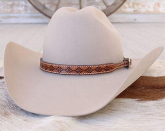 May include: A light beige cowboy hat with a brown leather band featuring a decorative pattern. The hat has a classic western design with a wide brim and a tall crown. The leather band is secured with a silver-colored buckle.